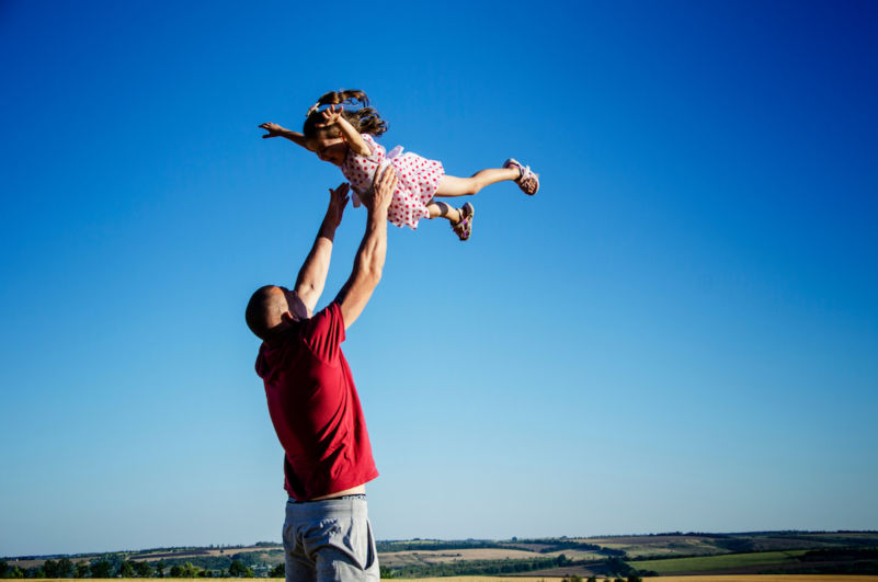 A father playfully throws his young daughter into the air while she laughs, symbolising trust and joy.