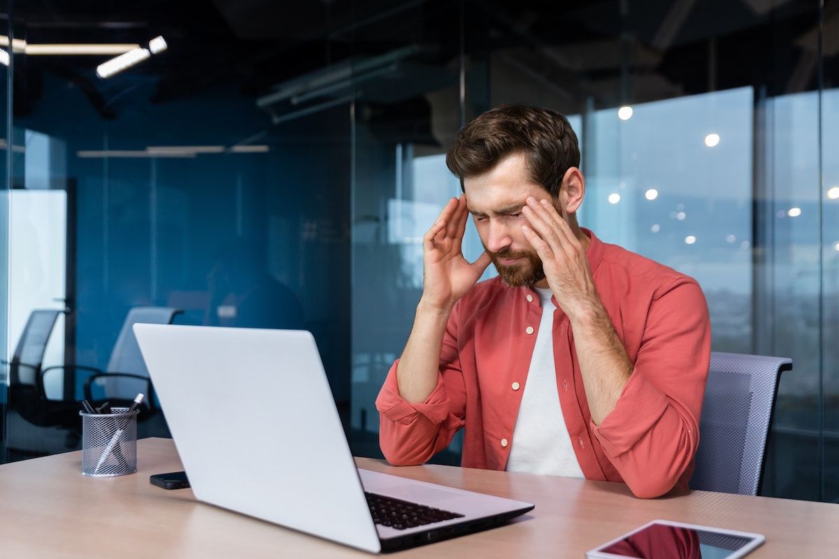 Stress and headache at work. A young businessman in a red shirt is sitting at the desk in the office, holding his head, close eyes. He feels bad, worries about problems, a project, overtime,