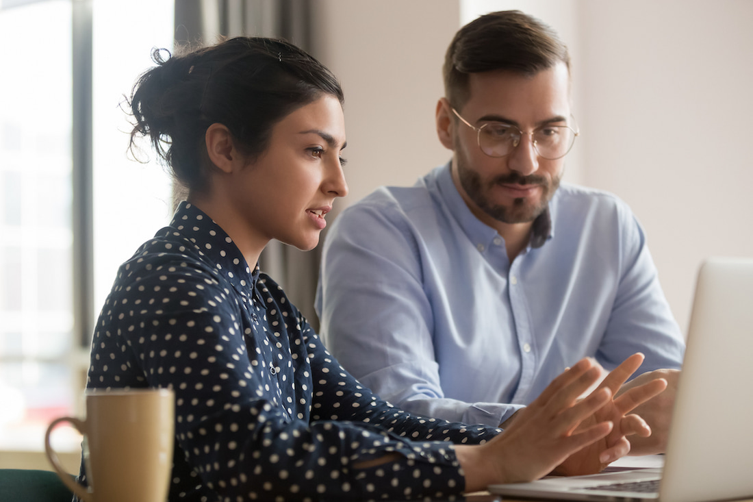 Focused male intern listening to serious indian business woman mentor teacher explaining online strategy looking at laptop computer teach trainee training new worker learning new skill at workplace